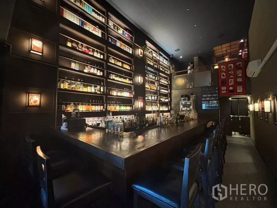 Shophouse for rent in Watthana, Bangkok - Dimly lit bar with a long wooden counter and tall illuminated liquor shelves with a mezzanine above.