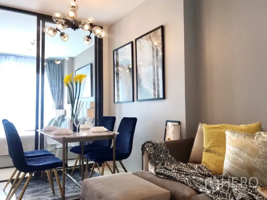 Condominium for rent or sale in Pathum Wan, Bangkok - Modern living and dining area with blue upholstered chairs, glass partition to the bedroom, wall art and statement chandelier.