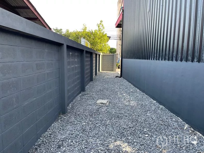 Warehouse for rent in Bang Kapi, Bangkok - Gravel side passage between a tall concrete boundary wall and corrugated metal warehouse wall.