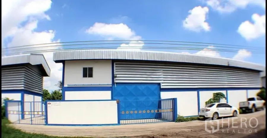 Factory for rent in Pak Kret, Nonthaburi - Blue-and-white factory exterior with corrugated metal roof, wide sliding gate and roadside parking.