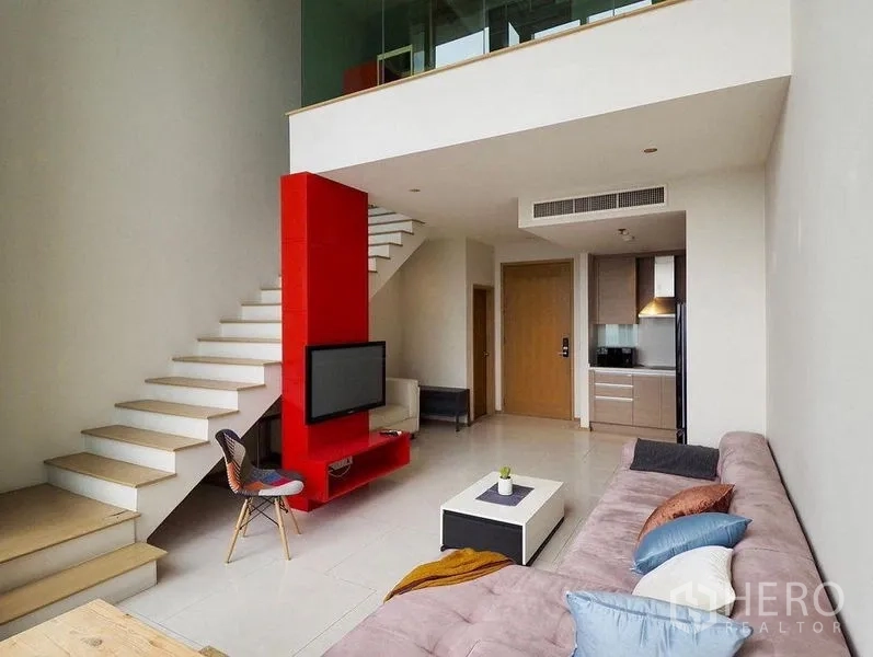 Condominium for rent in Khlong Toei, Bangkok - Double-height living area with sectional sofa, mezzanine and bold red feature column.