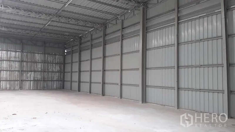 Warehouse for rent in Lat Phrao, Bangkok - Side view of empty warehouse showing metal cladding walls, roof trusses and wide open concrete floor for storage.