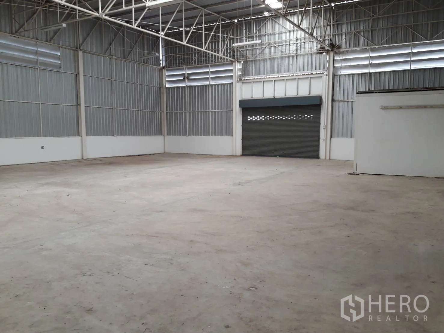 Warehouse for rent or sale in Khlong Luang, Pathum Thani - Spacious warehouse interior with concrete floor, high steel truss roof and a roller shutter door.