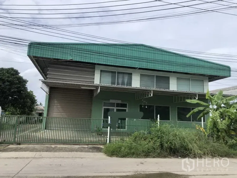 Factory for rent or sale in Samut Prakan - Front elevation of a two-storey office and warehouse building with green fence and large roller door.