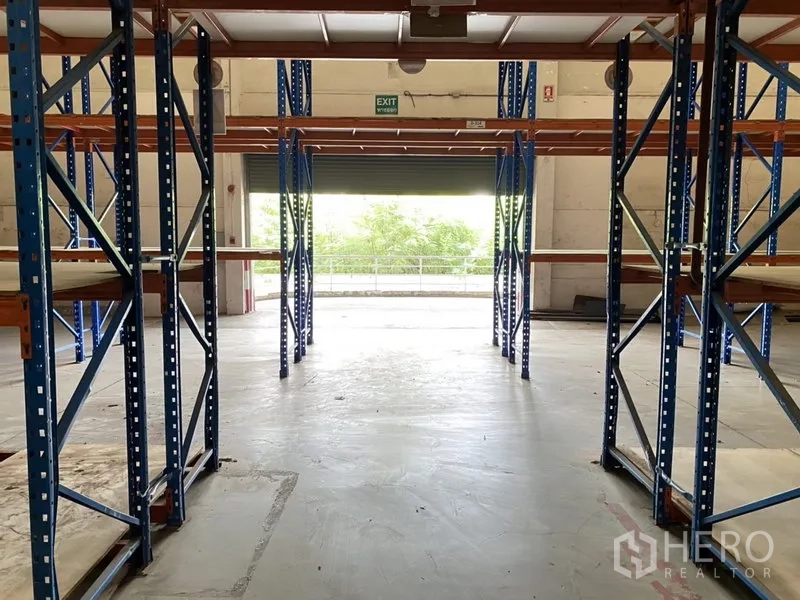 Warehouse for rent in Bang Phli, Samut Prakan - Warehouse aisle facing a roller shutter door with shelving and smooth concrete floor.