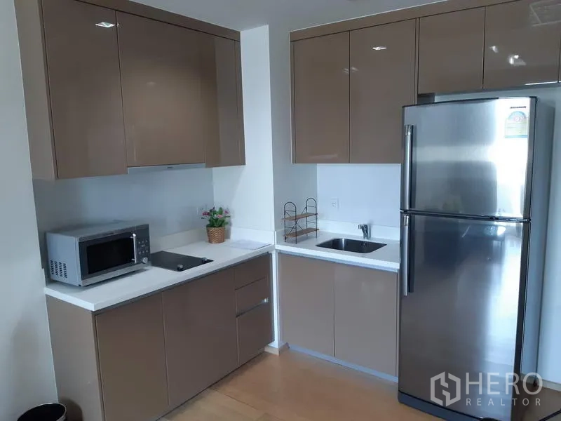Condominium for rent in Khlong Toei, Bangkok - Modern corner kitchen with glossy taupe cabinets, white countertops, stainless refrigerator and microwave.