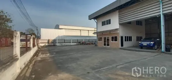 Warehouse for rent in Khlong Luang, Pathum Thani - Spacious concrete yard with an attached two-storey office beside the warehouse loading bays, suitable for truck parking.