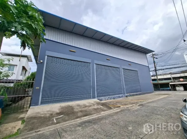 Warehouse for rent in Suan Luang, Bangkok - Side exterior showing three large metal roller shutter bays with a wide loading apron for easy truck access.