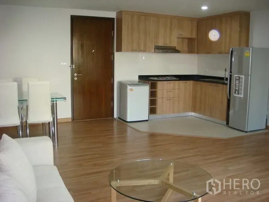 Apartment for rent in Khlong Toei, Bangkok - Open-plan living area with wood-tone L-shaped kitchen, refrigerator and dining set in an Asok apartment.