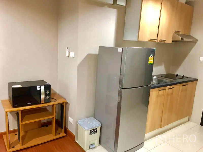 Condominium for rent in Huai Khwang, Bangkok - Compact kitchenette with refrigerator, microwave on a stand, sink, and wood cabinets.