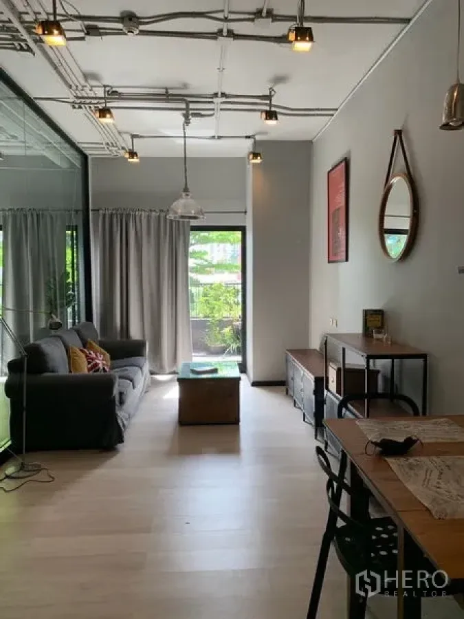 Condominium for sale in Watthana, Bangkok - Bright living room with grey sofa, pendant lights, light wood flooring, and glass door opening to a leafy balcony.