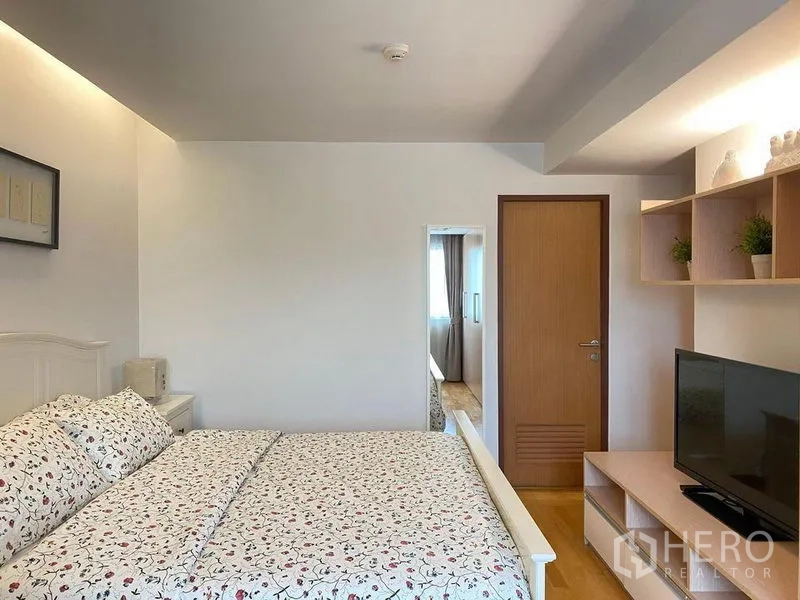 Condominium for sale in Phra Khanong, Bangkok - Bedroom view with floral bedspread, TV console, and open wall shelves.