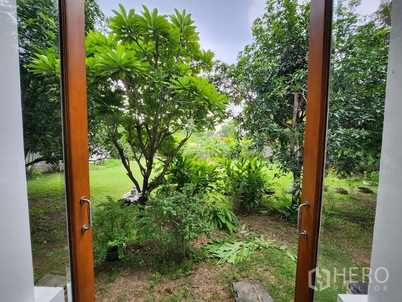 Detached House for rent in Prawet, Bangkok - Garden view framed by wooden French doors, showing mature tropical trees and lawn.