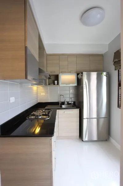 Townhouse for rent in Phra Khanong, Bangkok - Compact L-shaped kitchen with gas cooktop, wood cabinetry and stainless refrigerator.