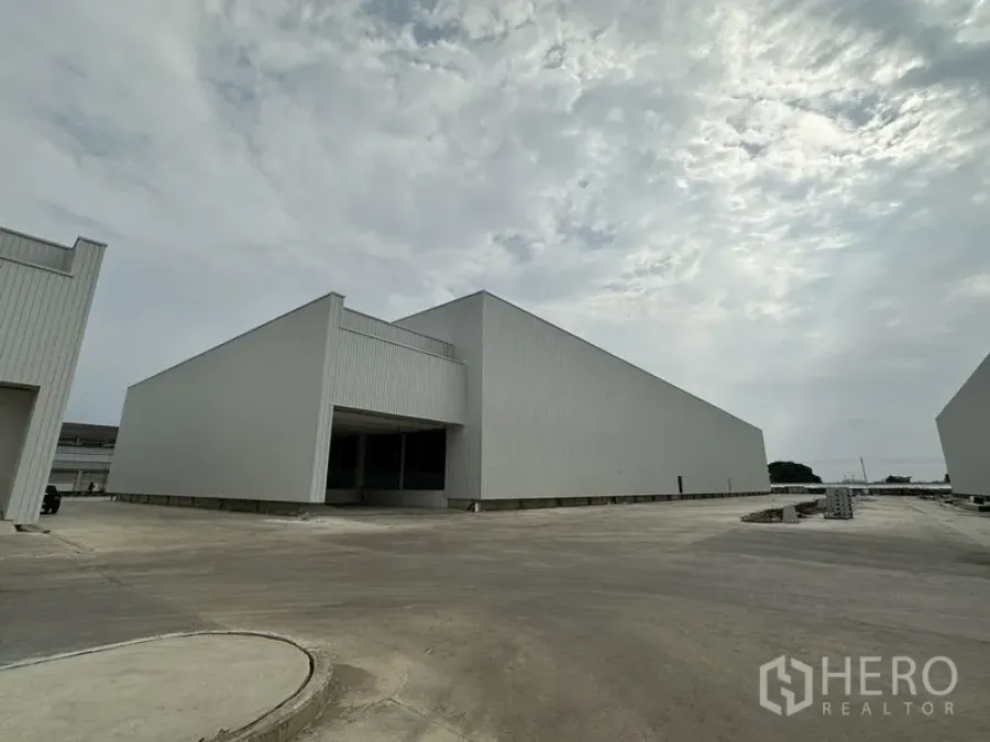 Factory for rent in Pathum Thani - New grey-clad factory buildings with a wide concrete driveway in an industrial park under a cloudy sky in Pathum Thani.