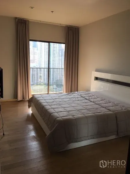 Condominium for rent in Bangkok - Sunlit bedroom with wood flooring, a modern white bed and a large window opening to a city-view balcony.