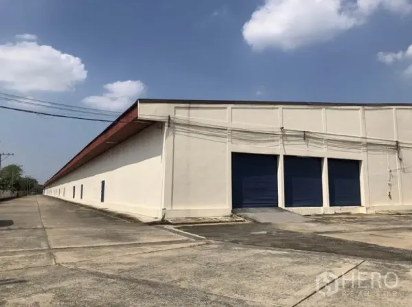 Warehouse for rent in Lam Luk Ka, Pathum Thani - Side elevation showing multiple dock-high blue shutters and a long driveway.