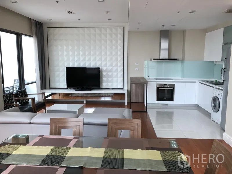 Condominium for rent or sale in Khlong Toei, Bangkok - Open-plan living room with TV feature wall, modern white kitchen and wood floors.