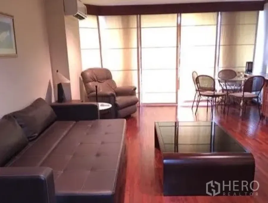 Condominium for rent or sale in Watthana, Bangkok - Living room with leather sofa, glass coffee table and dining nook by windows.