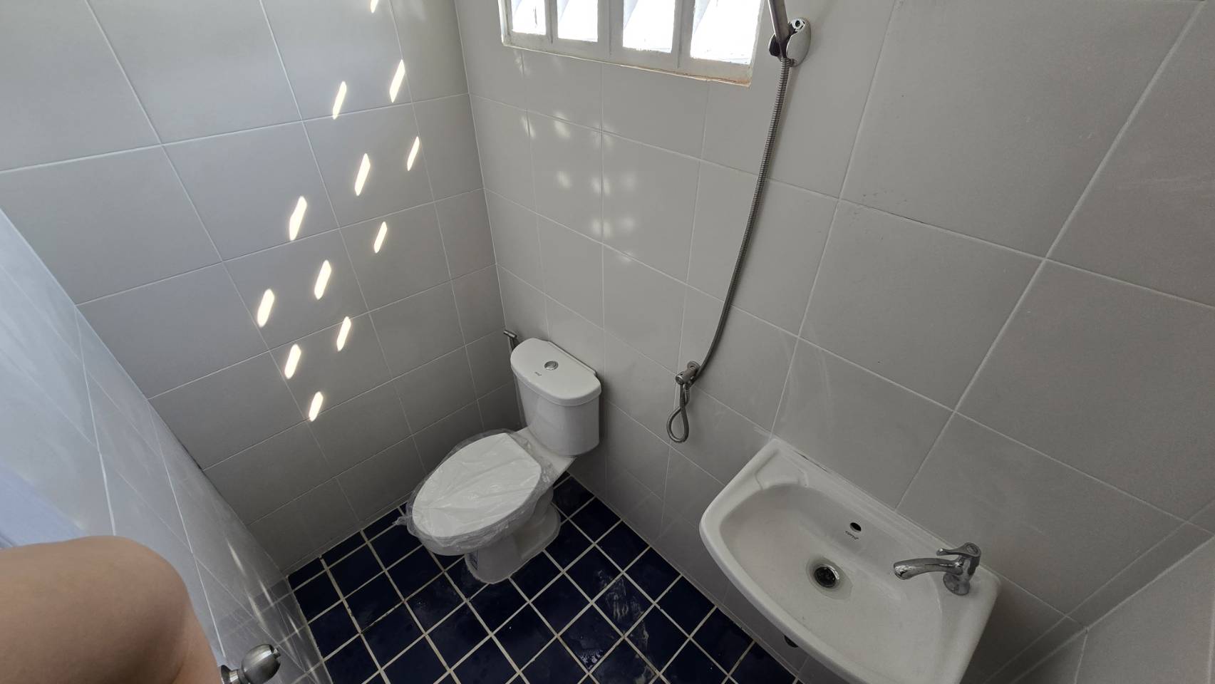 Warehouse for rent in Bang Phli, Samut Prakan - Clean tiled restroom with toilet, handheld bidet spray, sink, and a small window for ventilation.