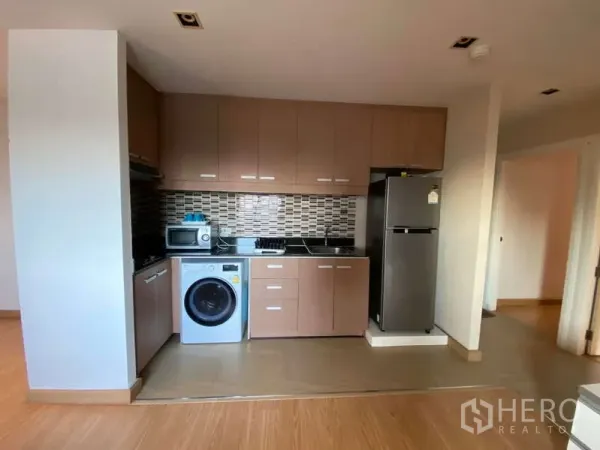 Condominium for rent in Khlong Toei, Bangkok - Compact fitted kitchen with brown cabinets, mosaic tile backsplash, cooktop, microwave, fridge and washing machine.