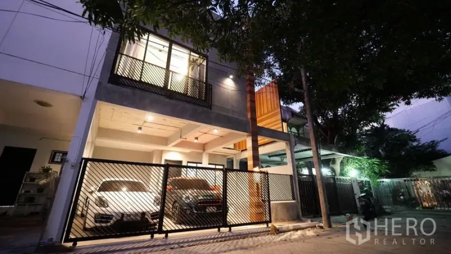 Townhouse for rent in Khlong Toei, Bangkok - Modern townhouse exterior at dusk with gated entrance and covered parking for two cars on a tree-lined lane.