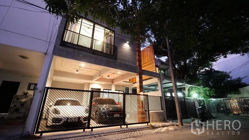 Townhouse for rent in Khlong Toei, Bangkok - Modern townhouse exterior at dusk with gated entrance and covered parking for two cars on a tree-lined lane.