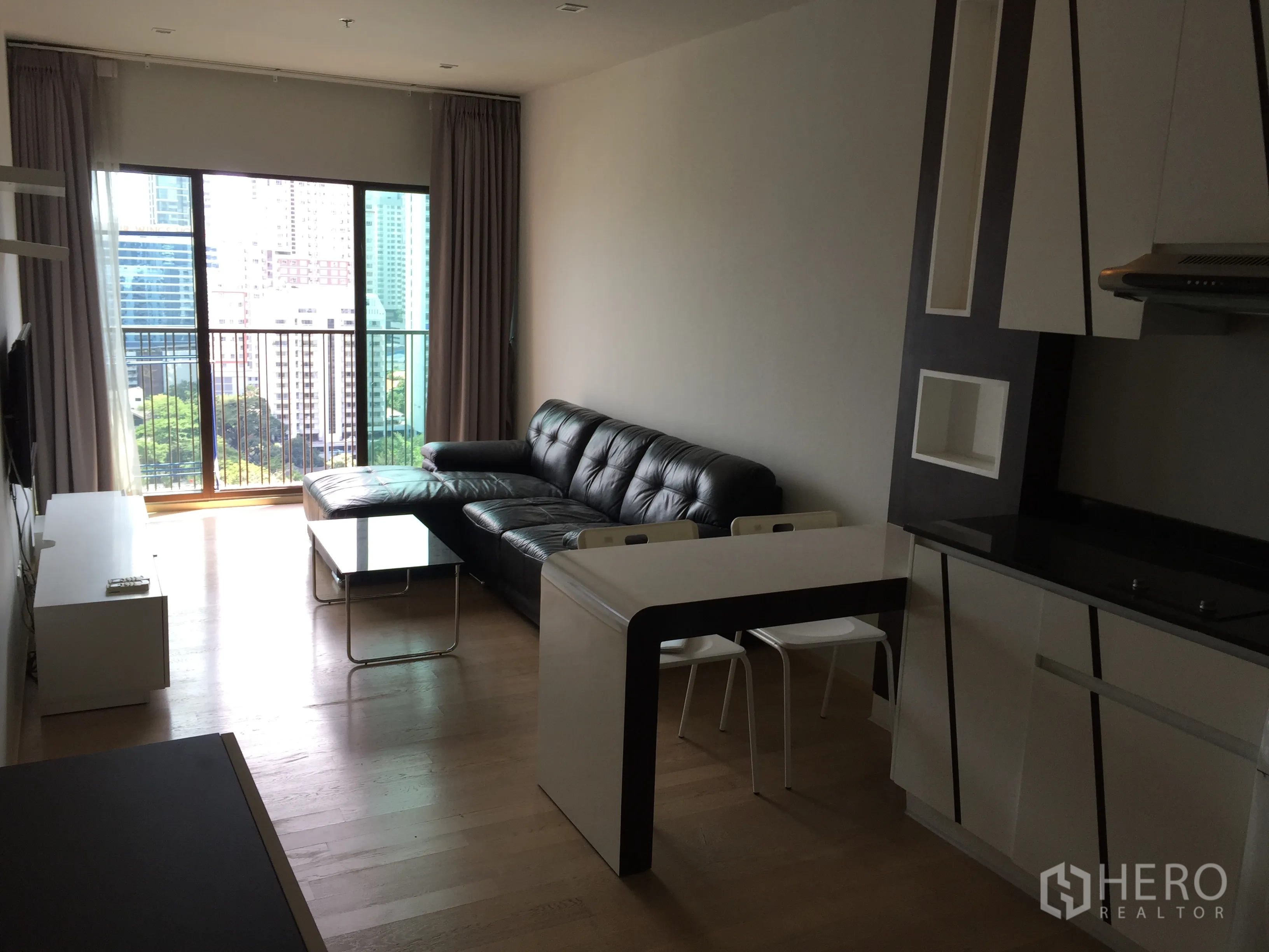 Condominium for rent in Bangkok - Open-plan living room with black leather sectional, balcony city views and a compact fitted kitchen.