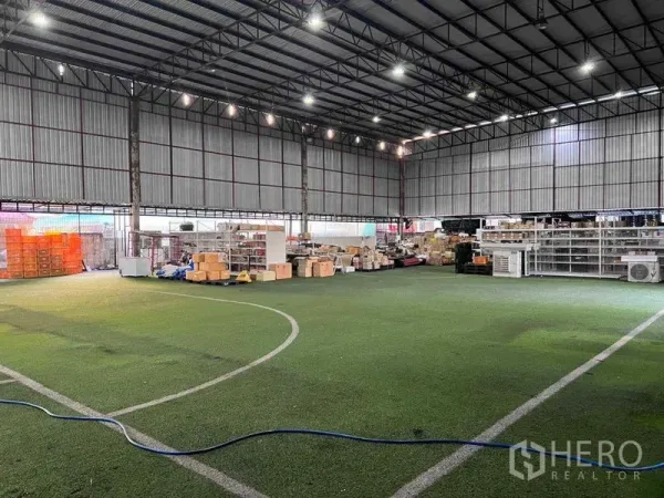 Warehouse for rent in Lat Phrao, Bangkok - High-clearance steel-frame warehouse interior with artificial turf flooring, bright LED lights, and storage racks with boxes.