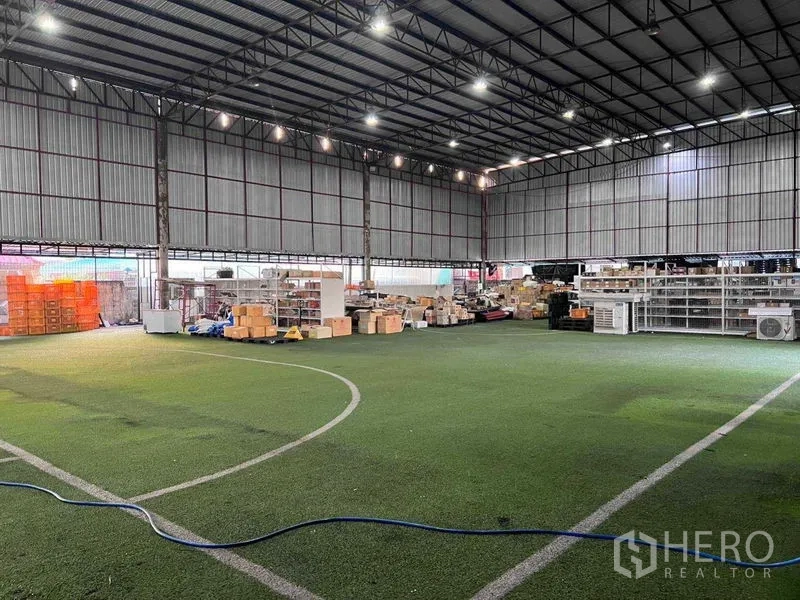 Warehouse for rent in Lat Phrao, Bangkok - High-clearance steel-frame warehouse interior with artificial turf flooring, bright LED lights, and storage racks with boxes.