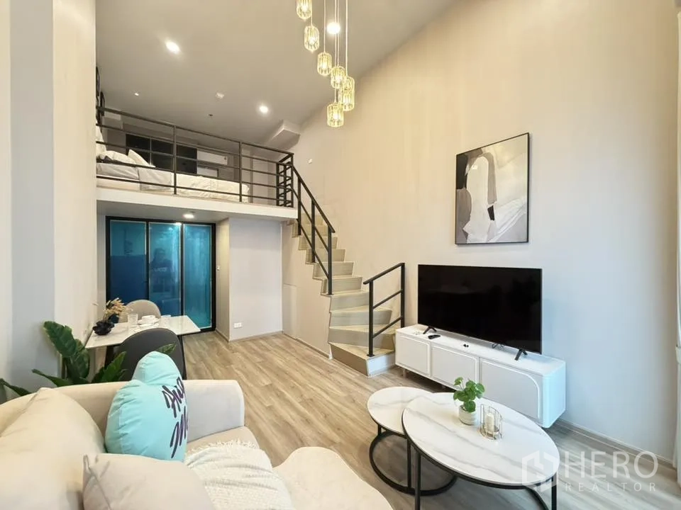 Condominium for rent in Khlong Toei, Bangkok - Loft-style living room with mezzanine bedroom, staircase, sofa, TV console, dining nook, and pendant lights in a modern condo.