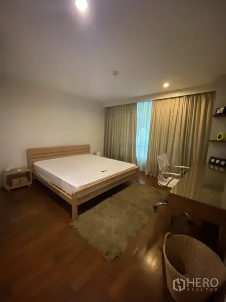 Condominium for rent or sale in Khlong Toei, Bangkok - Spacious bedroom with large bed, study desk and chair, soft rug on wooden floors, and wide window with curtains.