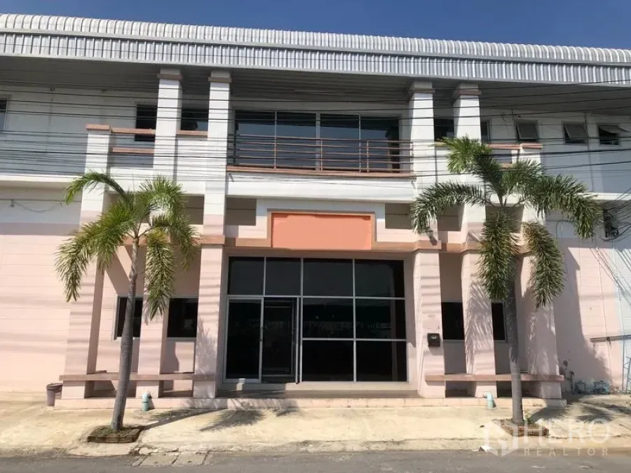 Factory for rent in Pak Kret, Nonthaburi - Two-storey factory exterior with glass entrance, balcony railings and palm trees in Pak Kret.