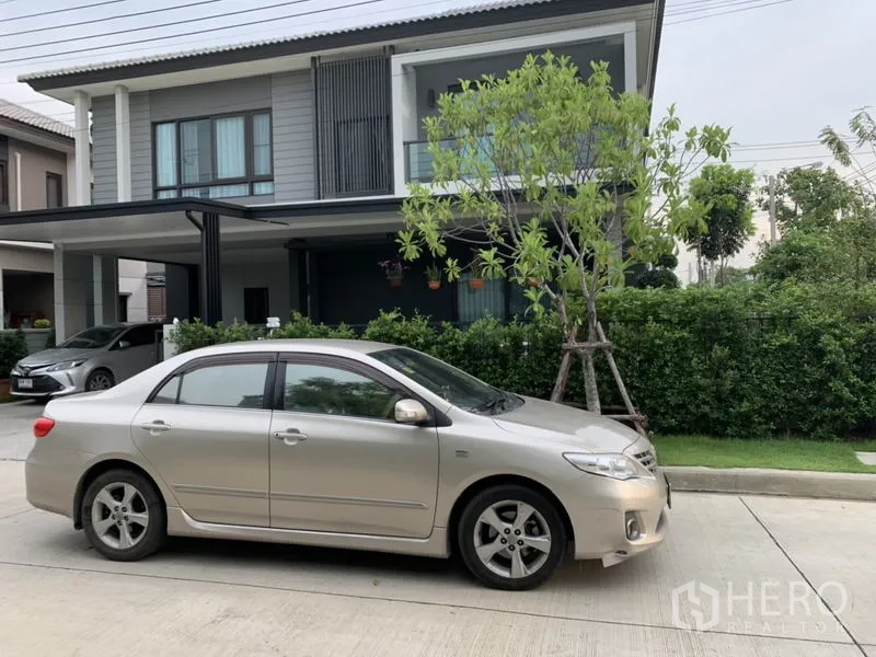 Detached House for rent or sale in Samut Prakan - Modern two-storey detached house facade with carport, balcony and hedge-lined frontage.