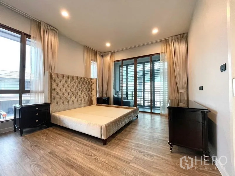 Detached House for sale in Bang Kapi, Bangkok - Spacious master bedroom with large windows, wood flooring, tufted headboard frame and doors to a balcony.