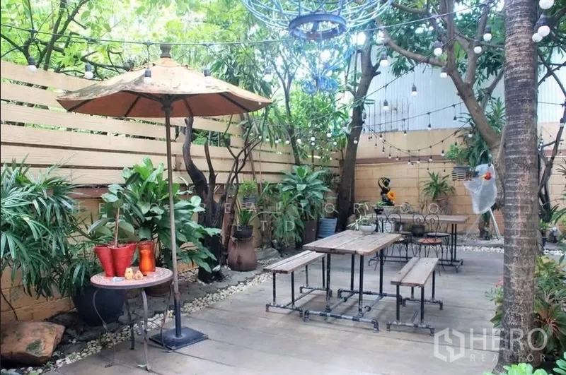 Detached House for rent in Watthana, Bangkok - Leafy outdoor seating area with benches, parasol and decorative lights on a walled patio.