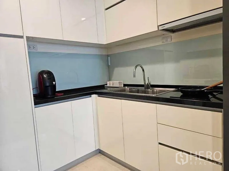 Condominium for rent or sale in Khlong Toei, Bangkok - Modern L-shaped kitchen with glossy white cabinets, black countertop, glass backsplash, sink and cooktop.
