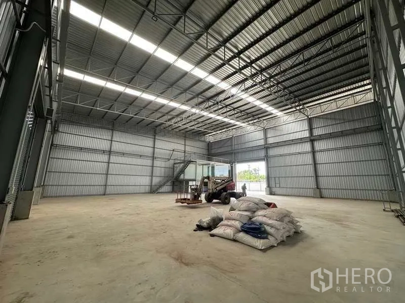 Factory for rent in Wang Noi, Phra Nakhon Si Ayutthaya - Large storage area with high truss roof, open shutter, and materials placed on the floor.