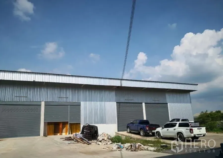 Warehouse for rent in Khlong Luang, Pathum Thani - Exterior of metal-clad warehouse units with gray roller shutters, parking area and pickup trucks under a blue sky in Khlong Luang.