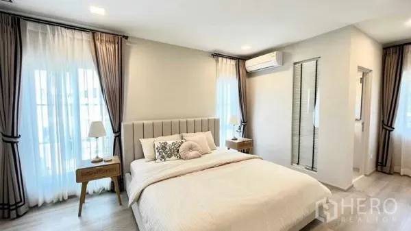 บ้าน ให้เช่าหรือขายในPrawet, Bangkok - ห้องนอนใหญ่สว่าง เตียงบุผ้า โต๊ะข้าง ม่านทึบโปร่ง และทางเข้าห้องน้ำ