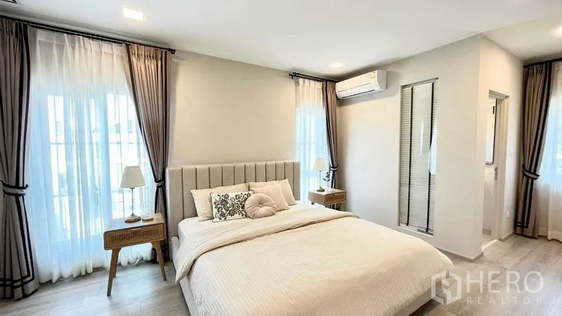 บ้าน ให้เช่าหรือขายในPrawet, Bangkok - ห้องนอนใหญ่สว่าง เตียงบุผ้า โต๊ะข้าง ม่านทึบโปร่ง และทางเข้าห้องน้ำ