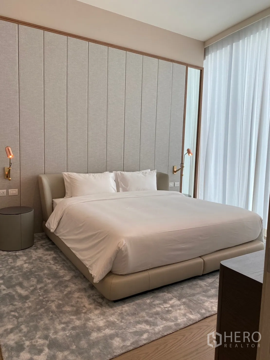 Condominium for rent in Pathum Wan, Bangkok - Elegant bedroom with upholstered bed, paneled headboard, floor-to-ceiling sheer curtains, soft rug, and warm bedside sconces.