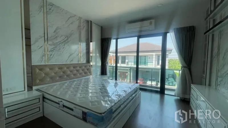 Detached House for rent or sale in Prawet, Bangkok - Primary bedroom with marble accent wall, built-in storage and sliding glass doors to a balcony.