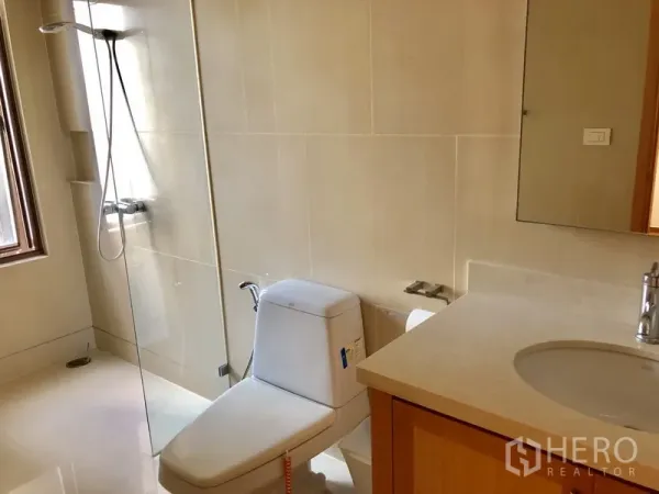 Condominium for rent in Khlong Toei, Bangkok - Modern bathroom with glass-enclosed shower, toilet and vanity in neutral stone finishes.