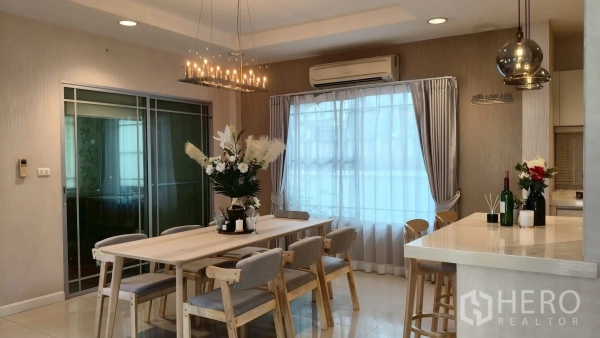Detached House for rent or sale in Mueang Samut Prakan, Samut Prakan - Dining area with long wooden table and six chairs under a chandelier beside the kitchen bar.