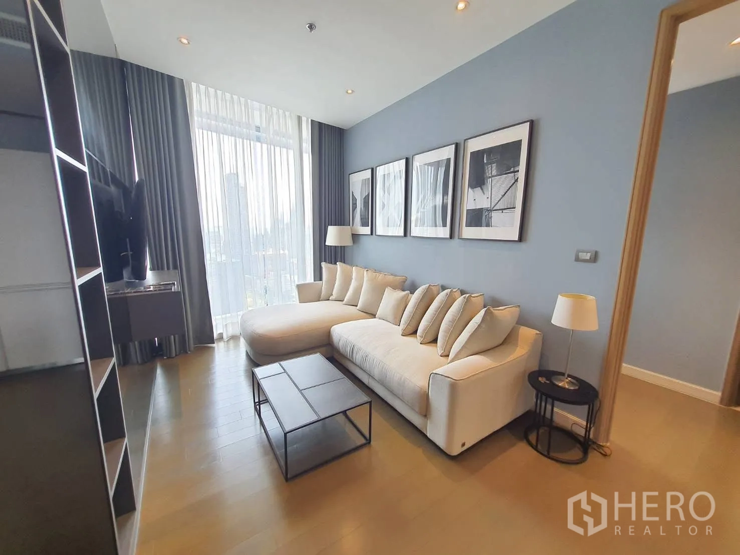 Condominium for rent in Pathum Wan, Bangkok - Modern living room with long white sofa, gallery art and floor-to-ceiling windows with sheer curtains and city view.