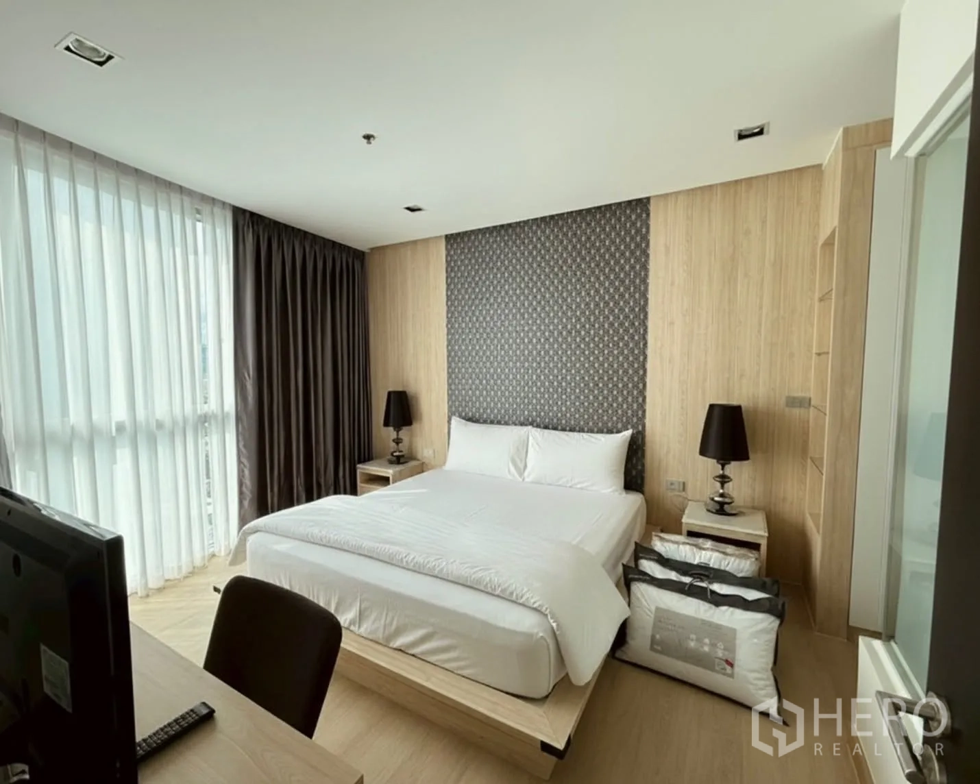 คอนโด for rent or sale in Watthana, Bangkok - ห้องนอนสว่างพร้อมเตียงขนาดใหญ่ หน้าต่างสูงจรดเพดาน ผนังลายไม้ และโคมไฟหัวเตียงข้างโต๊ะทำงาน