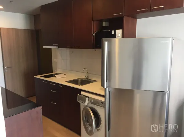 Condominium for rent in Bangkok - Compact kitchen with dark wood cabinets, white countertop, stainless refrigerator, and washing machine.