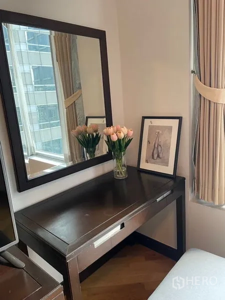 Condominium for rent in Pathum Wan, Bangkok - Vanity corner with mirror above wooden desk, vase of tulips and city window.
