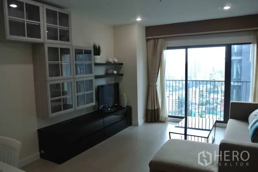 Condominium for rent in Khlong Toei, Bangkok - Bright condo living room with sofa, TV console, built-in shelves and balcony overlooking Bangkok city skyline.